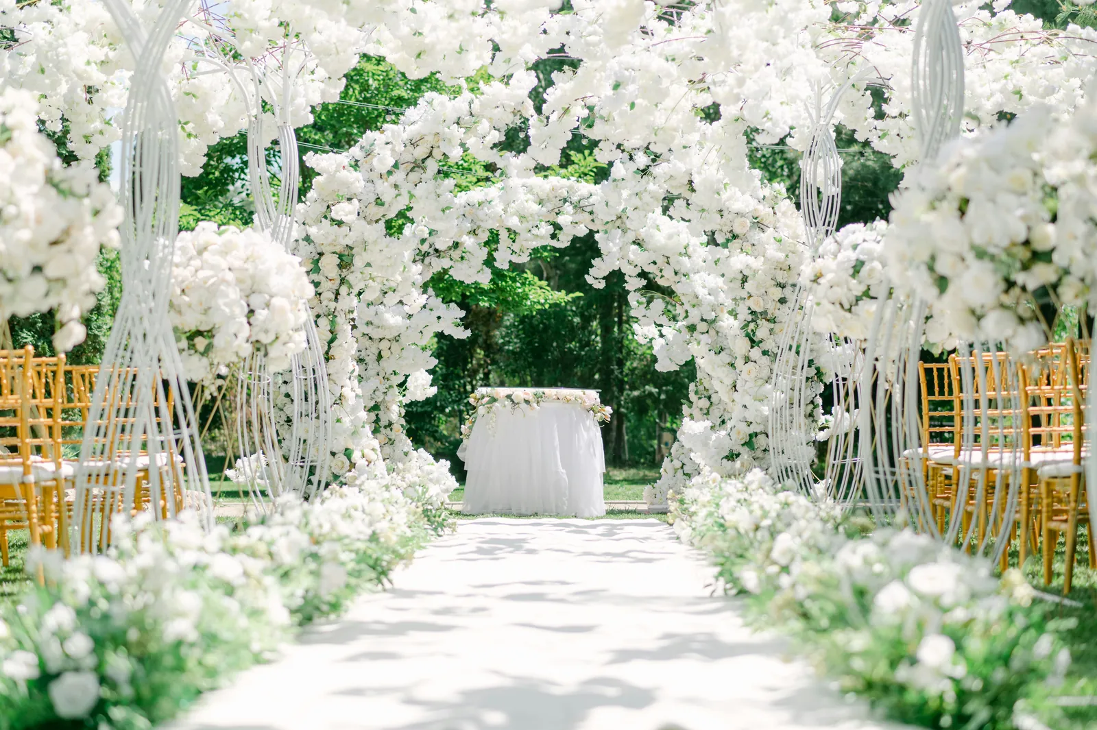 An outdoor ceremony aisle lined with lavish white floral arches and greenery.