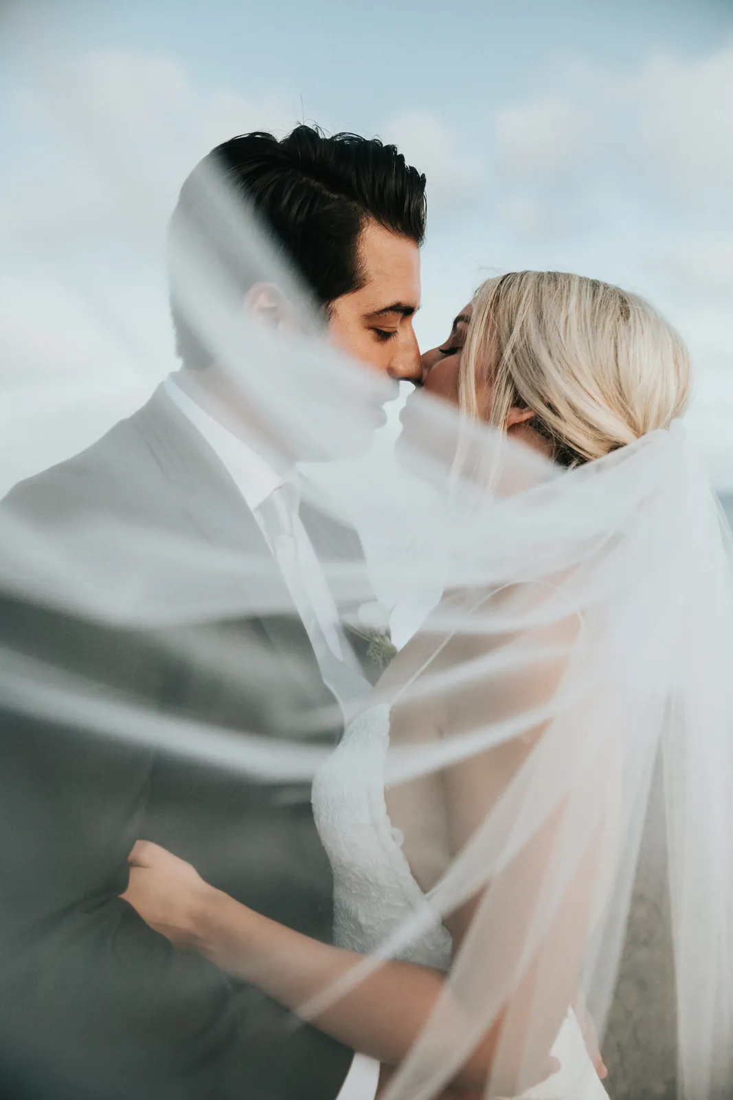 A bride and groom kissing beneath a flowing veil, backlit against a soft sky.