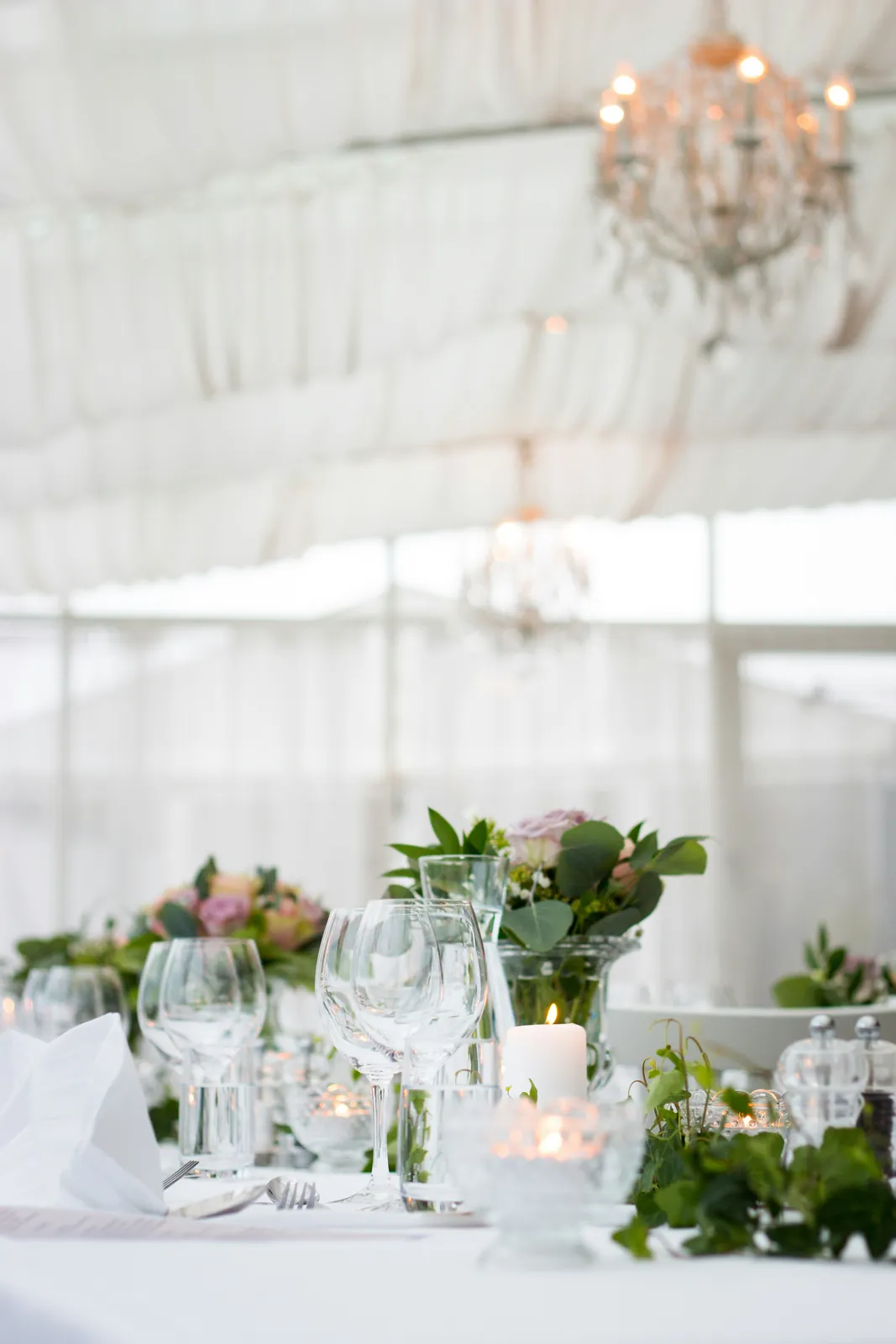 An elegant reception table set with glassware, candles and soft floral centerpieces beneath a chandelier.